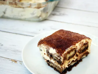 A slice of tiramisu on a plate dusted with cocoa powder, with the rest of the pan in the background