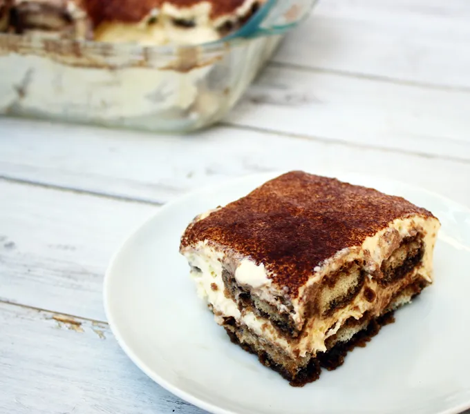 A slice of tiramisu on a plate dusted with cocoa powder, with the rest of the pan in the background
