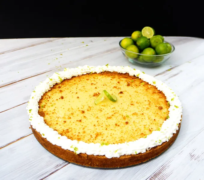 A whole key lime pie with a whipped cream border on a table, with a bowl of fresh key limes behind it against a black background