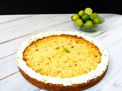 A whole key lime pie with a whipped cream border on a table, with a bowl of fresh key limes behind it against a black background
