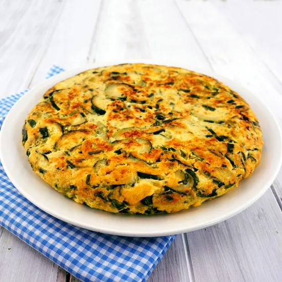 A whole zucchini frittata on a white plate, browned and studded with zucchini slices