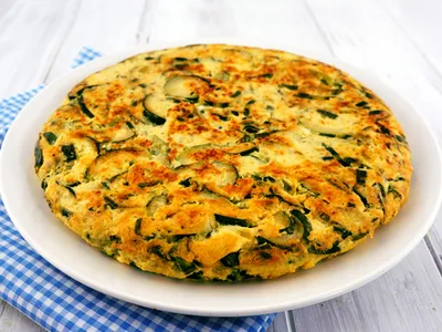 A whole zucchini frittata on a white plate, browned and studded with zucchini slices