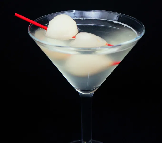 A martini glass filled with a clear lychee cocktail against a black background, garnished with two lychees on a red cocktail pick
