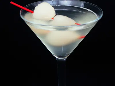A martini glass filled with a clear lychee cocktail against a black background, garnished with two lychees on a red cocktail pick