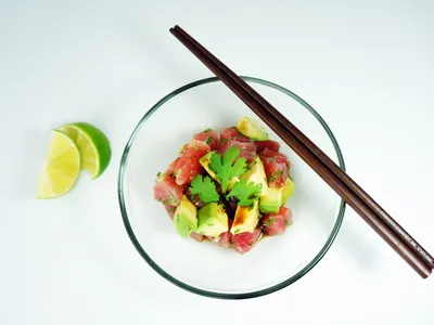 A glass bowl of diced tuna and avocado ceviche garnished with cilantro, chopsticks resting on top, lime wedges beside it