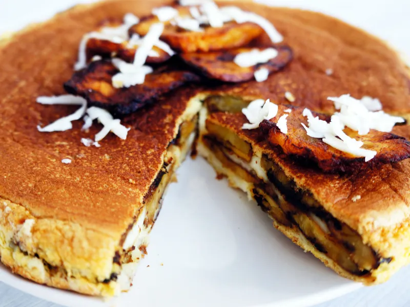 A plantain cake with a slice cut out, showing layers of plantain and cheese inside, topped with fried plantain slices and shredded cheese