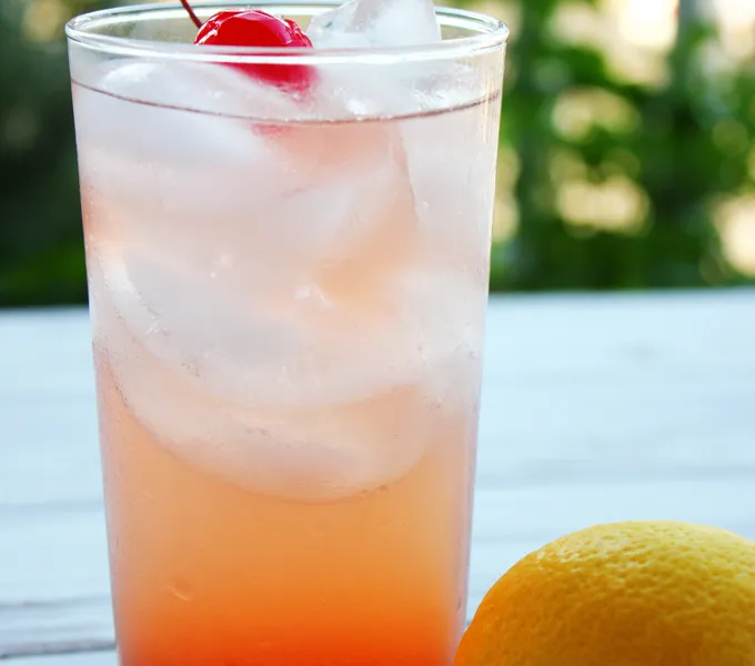 A tall glass of pink-orange lemonade with ice and a maraschino cherry, next to a whole lemon