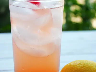 A tall glass of pink-orange lemonade with ice and a maraschino cherry, next to a whole lemon