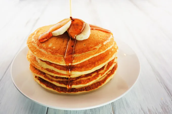 A tall stack of buttermilk pancakes topped with banana slices and syrup being poured over the top