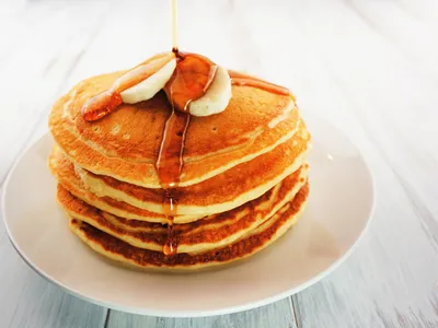 A tall stack of buttermilk pancakes topped with banana slices and syrup being poured over the top