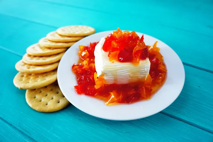 A block of cream cheese topped with red and orange pepper jelly on a plate, with crackers on the side