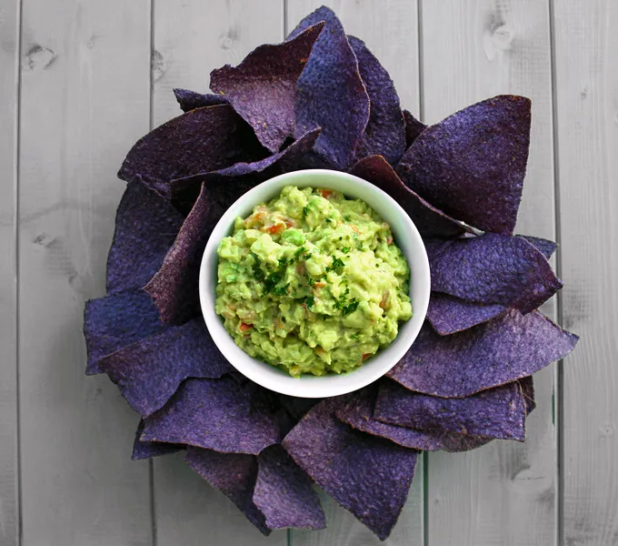 A bowl of guacamole surrounded by purple corn tortilla chips arranged like flower petals