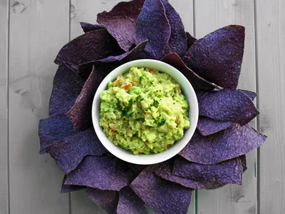 A bowl of guacamole surrounded by purple corn tortilla chips arranged like flower petals