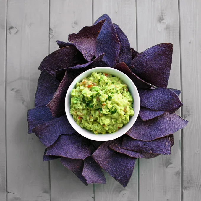 A bowl of guacamole surrounded by purple corn tortilla chips arranged like flower petals