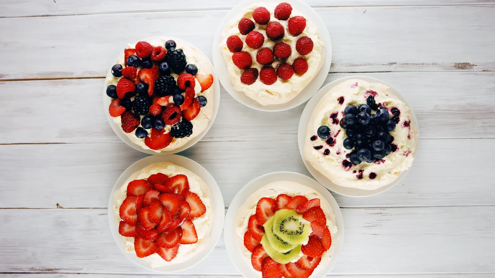 Five individual pavlovas topped with different fruit, arranged in a circle on a wooden table