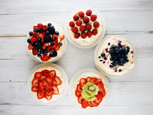 Five individual pavlovas arranged in a circle, each topped with a different fruit -- mixed berries, raspberries, blueberries, strawberries, and strawberry-kiwi
