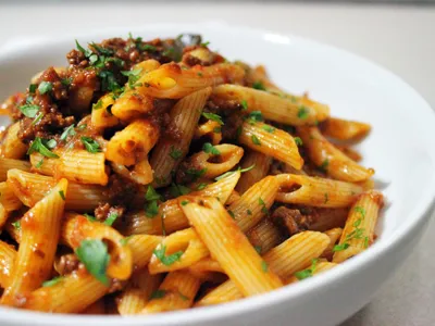 A bowl of penne pasta tossed in ragu sauce with ground beef and topped with chopped parsley