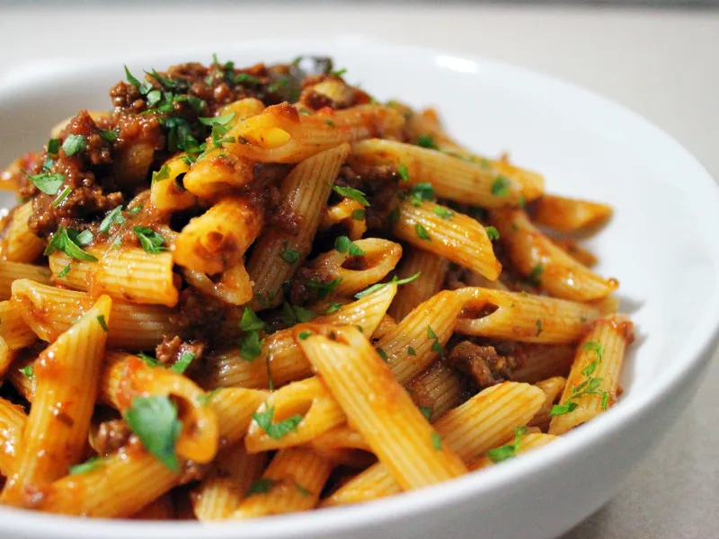 A bowl of penne pasta tossed in ragu sauce with ground beef and topped with chopped parsley