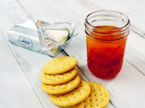 A jar of red pepper jelly next to a block of cream cheese and a stack of crackers