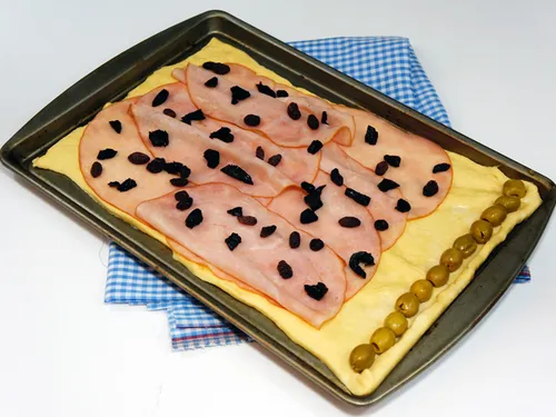Dough laid out on a baking sheet topped with ham, raisins, prunes, and a row of olives before rolling