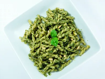 A square white bowl of gemelli pasta tossed in green basil pesto with pine nuts and a basil leaf garnish