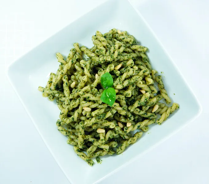 A square white bowl of gemelli pasta tossed in green basil pesto with pine nuts and a basil leaf garnish