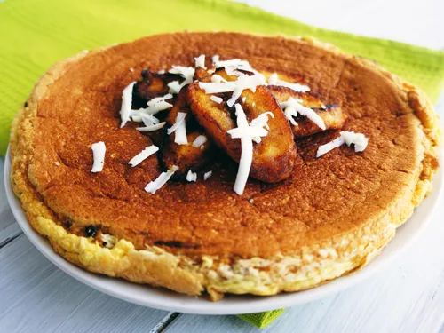 A whole, uncut plantain cake topped with fried plantain slices and shredded cheese on a green napkin