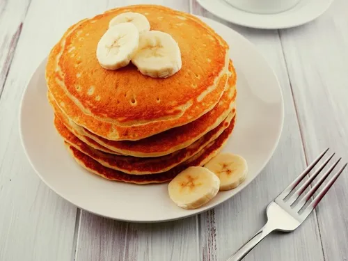 A stack of banana pancakes with a cup of coffee and a fork on the side