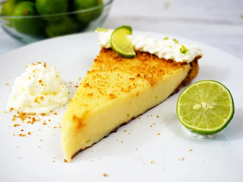 A slice of key lime pie on a plate with whipped cream and a lime wheel garnish, bowl of key limes in the background